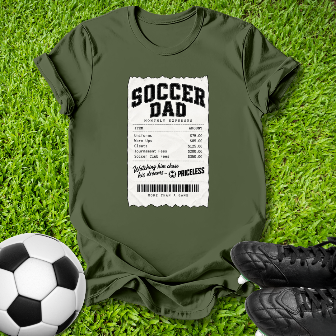 An olive green t-shirt laid flat on a green grass background, styled with a soccer ball on the left and black cleats on the right. The t-shirt features a white graphic print styled like a receipt with the text 'SOCCER DAD' and a humorous expense list.