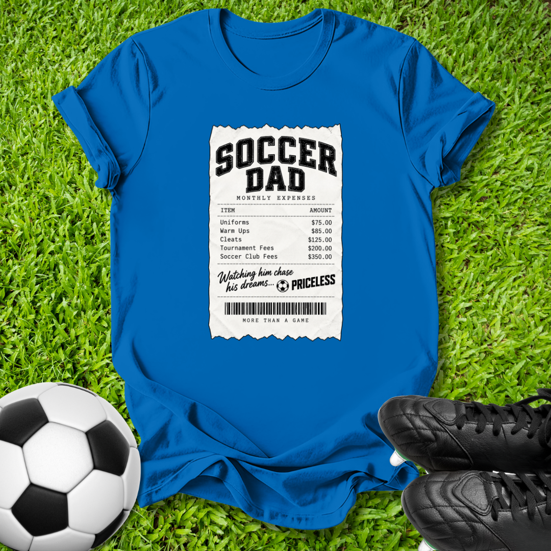 A bright blue graphic t-shirt laid flat on green grass, featuring a white and black 'Soccer Dad' receipt-style print on the front. A soccer ball is visible on the left, and black cleats are on the right, creating a themed sports flat-lay.