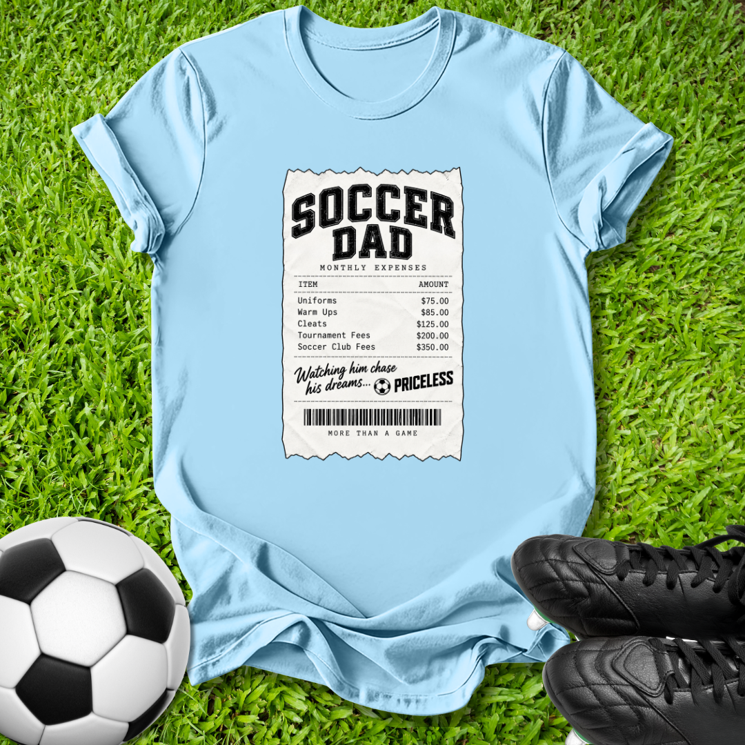 A light blue graphic t-shirt laid flat on green grass, styled with a soccer ball on the left and black soccer cleats on the right. The t-shirt features a white and black 'SOCCER MOM' receipt print graphic on the front.