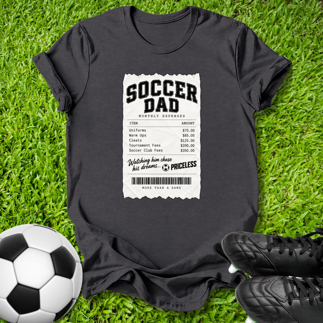 A dark charcoal gray t-shirt laid flat on green grass, featuring a white graphic print styled like a receipt with the text 'SOCCER DAD' and a fake barcodes. A soccer ball is on the left, and black cleats are on the right.