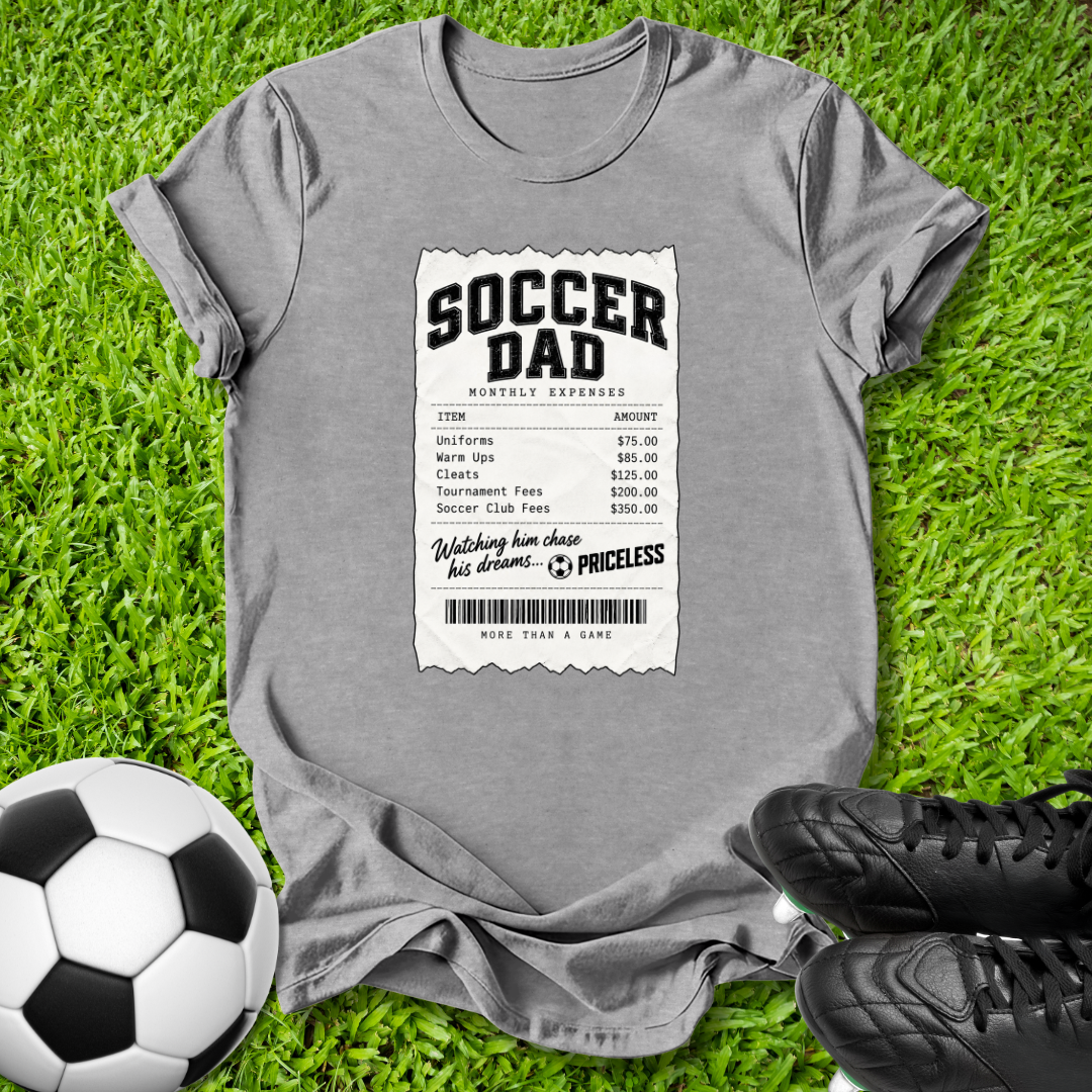 A gray short-sleeve t-shirt with a 'Soccer Dad' receipt graphic printed on the front, styled in a flat-lay on green grass with a soccer ball and black cleats.