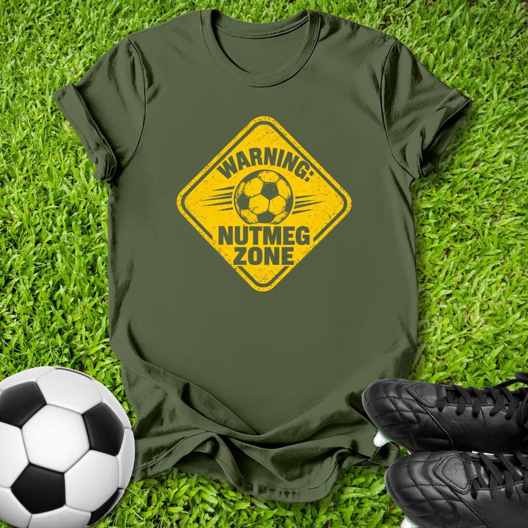 An olive green t-shirt with a yellow warning sign graphic that reads 'WARNING: NUTMEG ZONE' and features a soccer ball in the center. The shirt is laid flat on green grass, styled with a soccer ball and black cleats.
