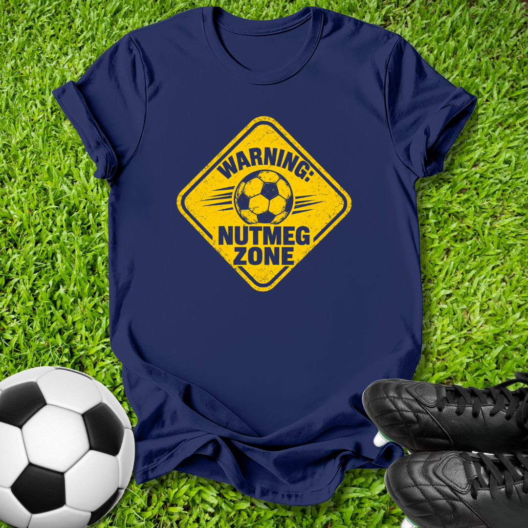 A navy blue short-sleeve t-shirt laid flat on green grass, featuring a yellow diamond-shaped warning sign graphic with a soccer ball and the text 'WARNING: NUTMEG ZONE'. A black-and-white soccer ball is at the bottom left, and black cleats are at the bottom right.