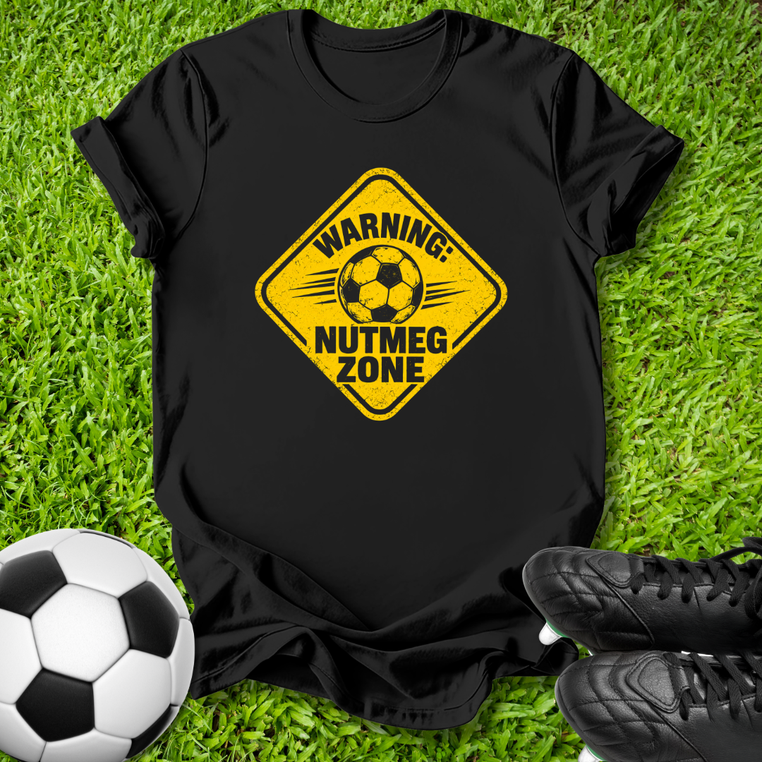 A black short-sleeve t-shirt laid flat on green grass, featuring a yellow diamond-shaped warning sign graphic with a soccer ball and the text 'WARNING: NUTMEG ZONE'. A soccer ball and black cleats are placed around it as props.