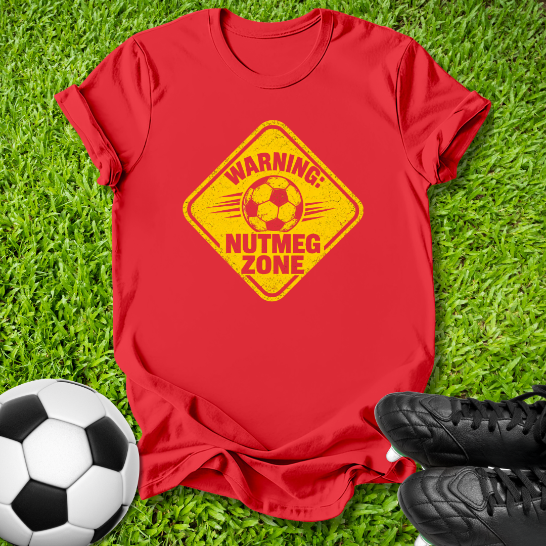 A red short-sleeve t-shirt laid flat on green grass, featuring a yellow diamond-shaped warning graphic with a soccer ball and the text 'WARNING: NUTMEG ZONE.' A black-and-white soccer ball appears at the lower left, and black cleats are at the lower right.