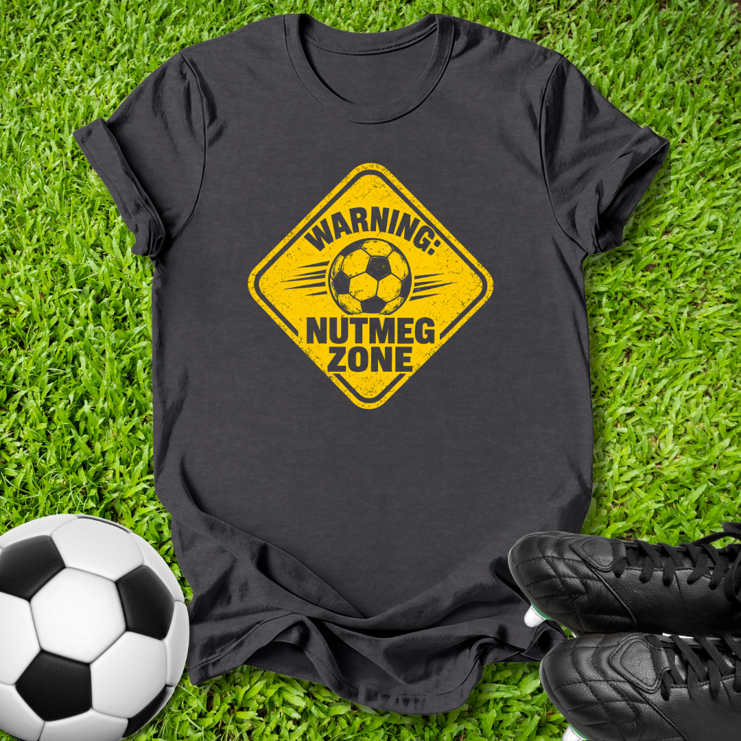 A black short-sleeve t-shirt laid flat on green grass, featuring a yellow diamond-shaped graphic with a soccer ball and the text 'WARNING: NUTMEG ZONE'. A black-and-white soccer ball is at the bottom left, and black cleats are at the bottom right.