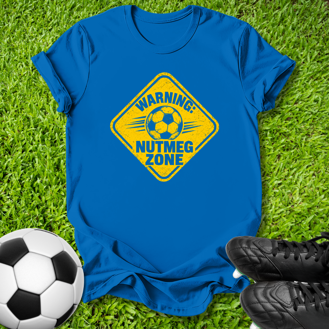 A royal blue short-sleeve t-shirt laid flat on green grass, featuring a yellow diamond-shaped graphic with a soccer ball and the text 'WARNING: NUTMEG ZONE'. A black-and-white soccer ball sits at the bottom left, and black cleats are at the bottom right.