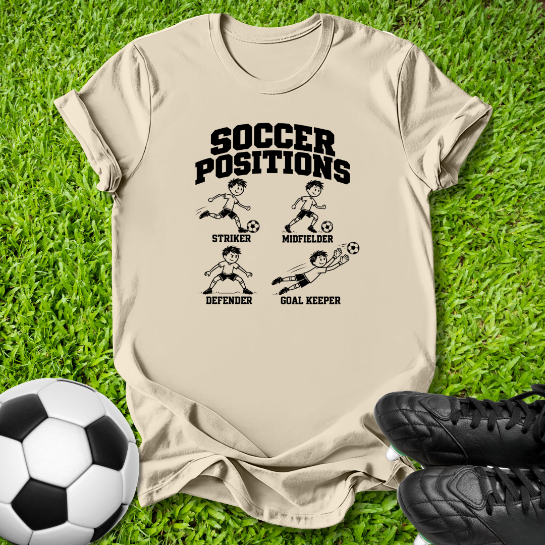 A beige graphic t-shirt laid flat on green grass, featuring a black 'SOCCER POSITIONS' print with illustrations of a striker, midfielder, defender, and goal keeper. A soccer ball is on the left and black cleats are on the right.