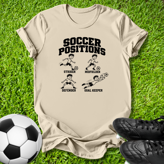 A beige graphic t-shirt laid flat on green grass, featuring a black 'SOCCER POSITIONS' print with illustrations of a striker, midfielder, defender, and goal keeper. A soccer ball is on the left and black cleats are on the right.