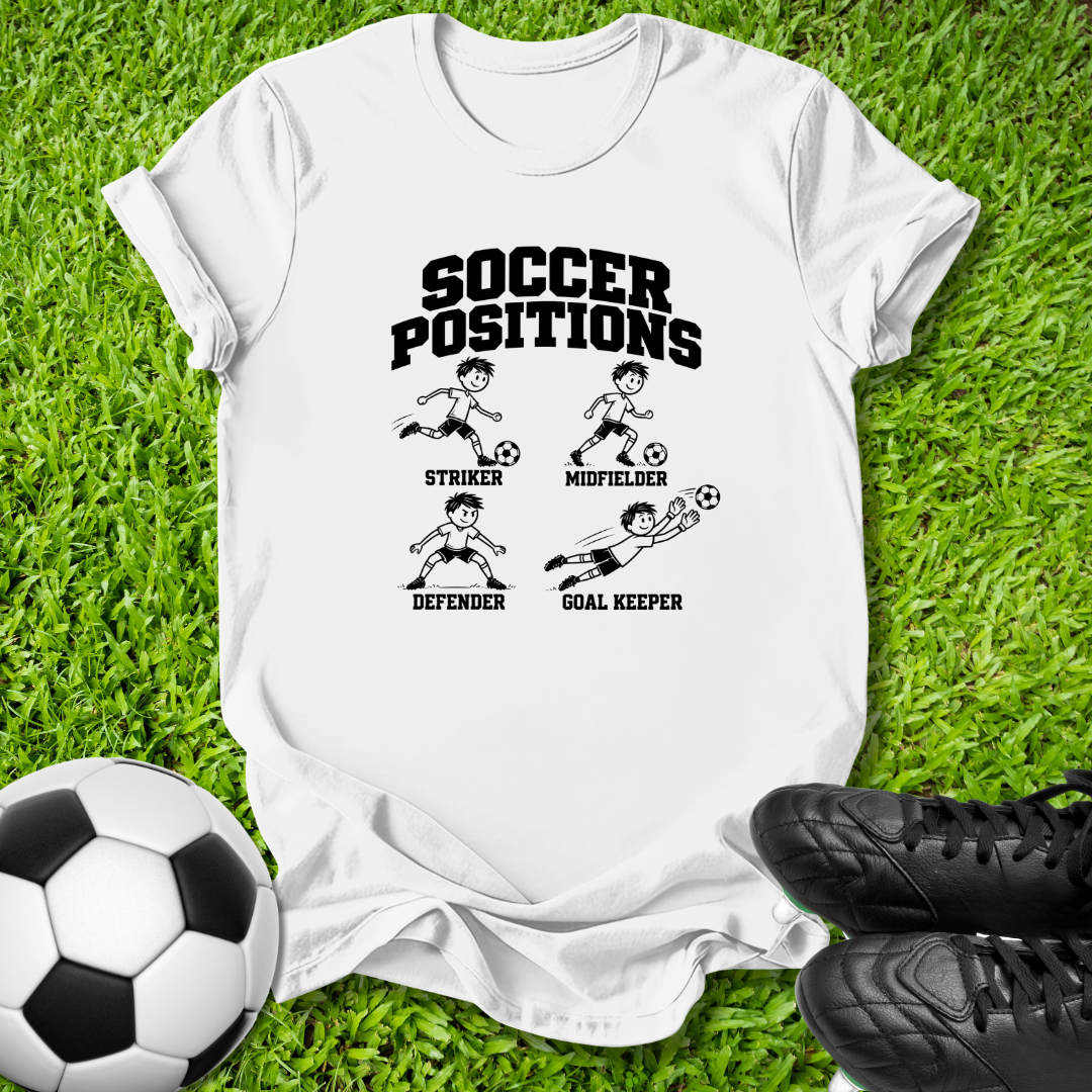 A white short-sleeve graphic T-shirt laid flat on green grass, with a soccer-themed print reading 'SOCCER POSITIONS' and simple illustrations of striker, midfielder, defender, and goal keeper. A black-and-white soccer ball appears at lower left, and black cleats are at lower right.