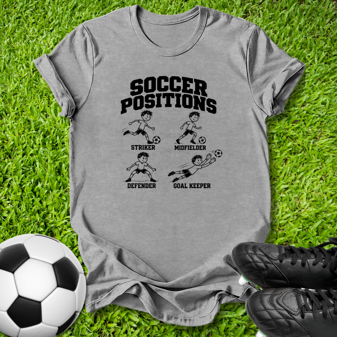 A heather gray short-sleeve t-shirt with a black 'SOCCER POSITIONS' graphic print, laid flat on green grass with a soccer ball and black cleats nearby.