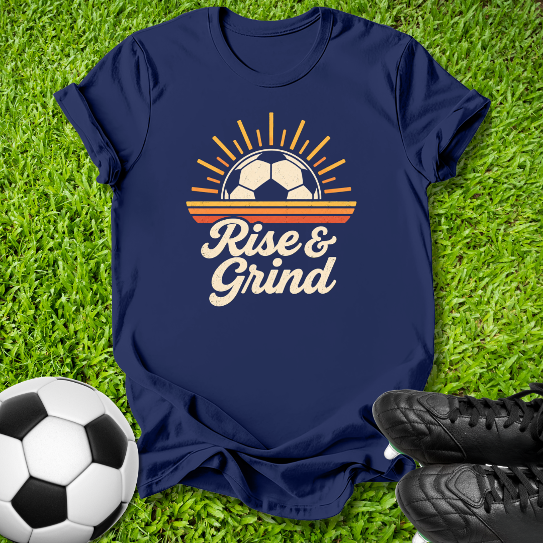 A navy blue graphic t-shirt laid flat on green grass, featuring a retro soccer design with a soccer ball in a sun and the text 'Rise & Grind'. A soccer ball and black cleats are styled around the shirt.