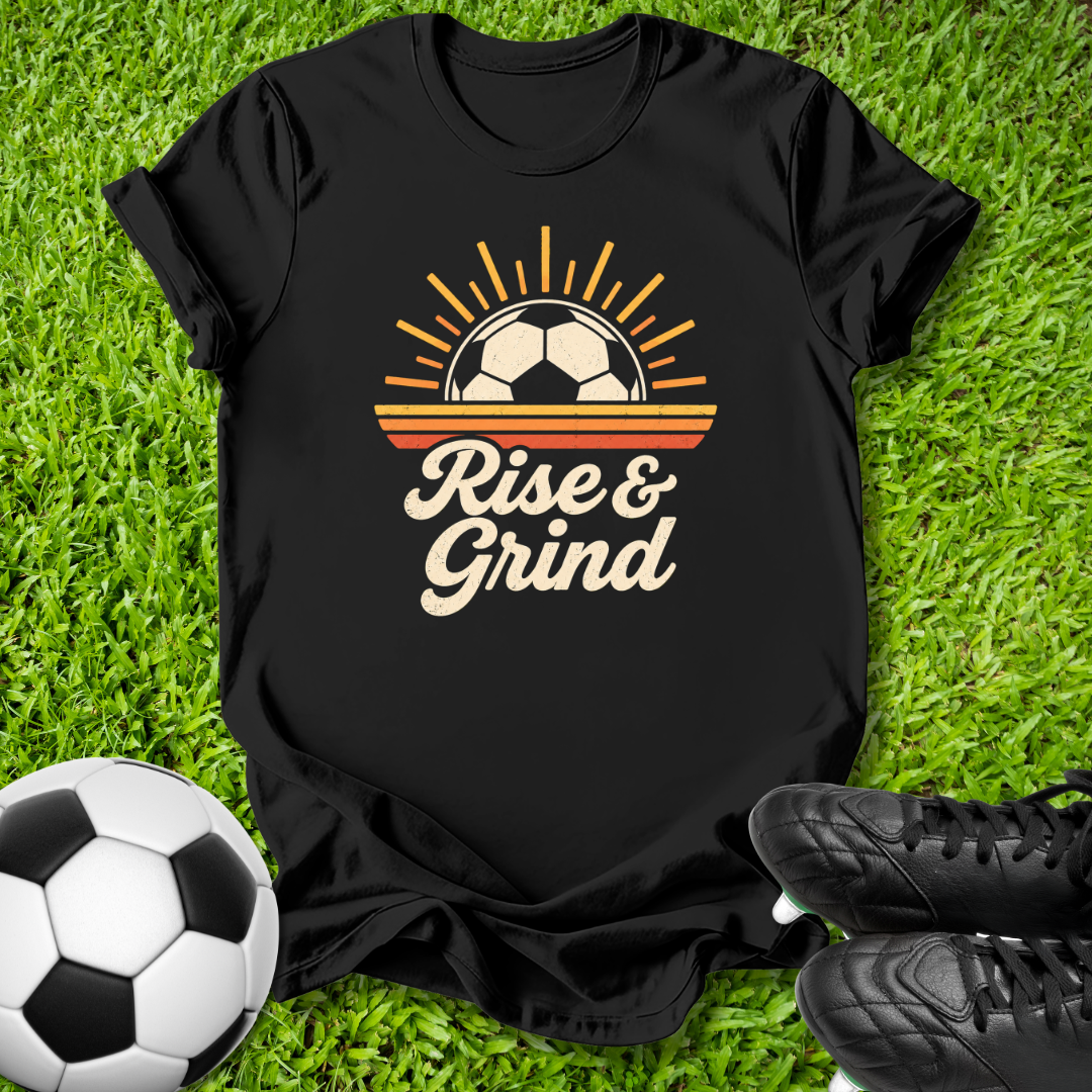 A black short-sleeve graphic T-shirt laid flat on green grass, with a retro soccer-themed print reading 'Rise & Grind' and a soccer ball graphic. A soccer ball and black cleats are placed around it as styling props.