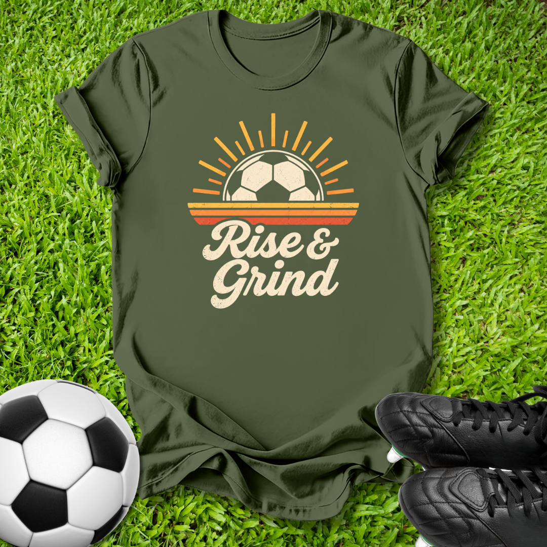 An olive green t-shirt laid flat on green grass, featuring a retro graphic of a soccer ball under a rising sun with the text 'Rise & Grind' in cream. A soccer ball and black cleats are styled around the shirt.