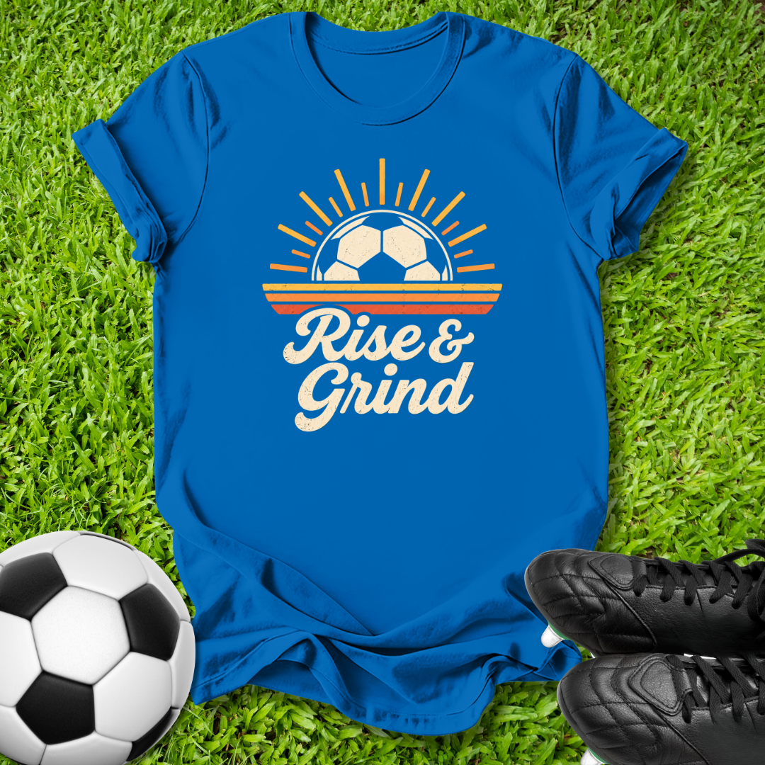 A royal blue graphic t-shirt laid flat on green grass, featuring a retro soccer-themed design with the text 'Rise & Grind'. A soccer ball is on the left and black cleats are on the bottom right.
