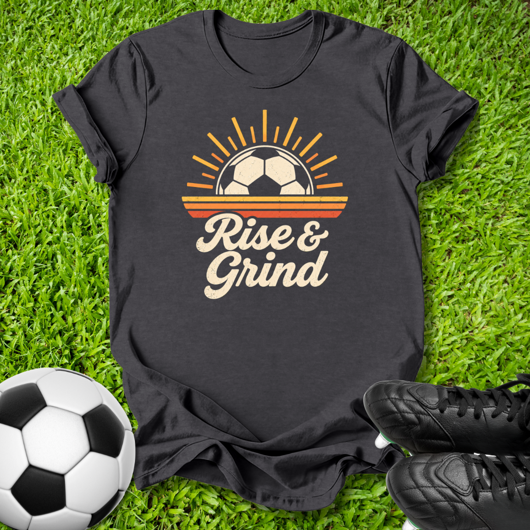 A black graphic t-shirt laid flat on green grass, featuring a retro soccer design with the text 'Rise & Grind'. A soccer ball is visible in the bottom-left corner and black cleats are in the bottom-right, creating a sports-themed flat-lay composition.