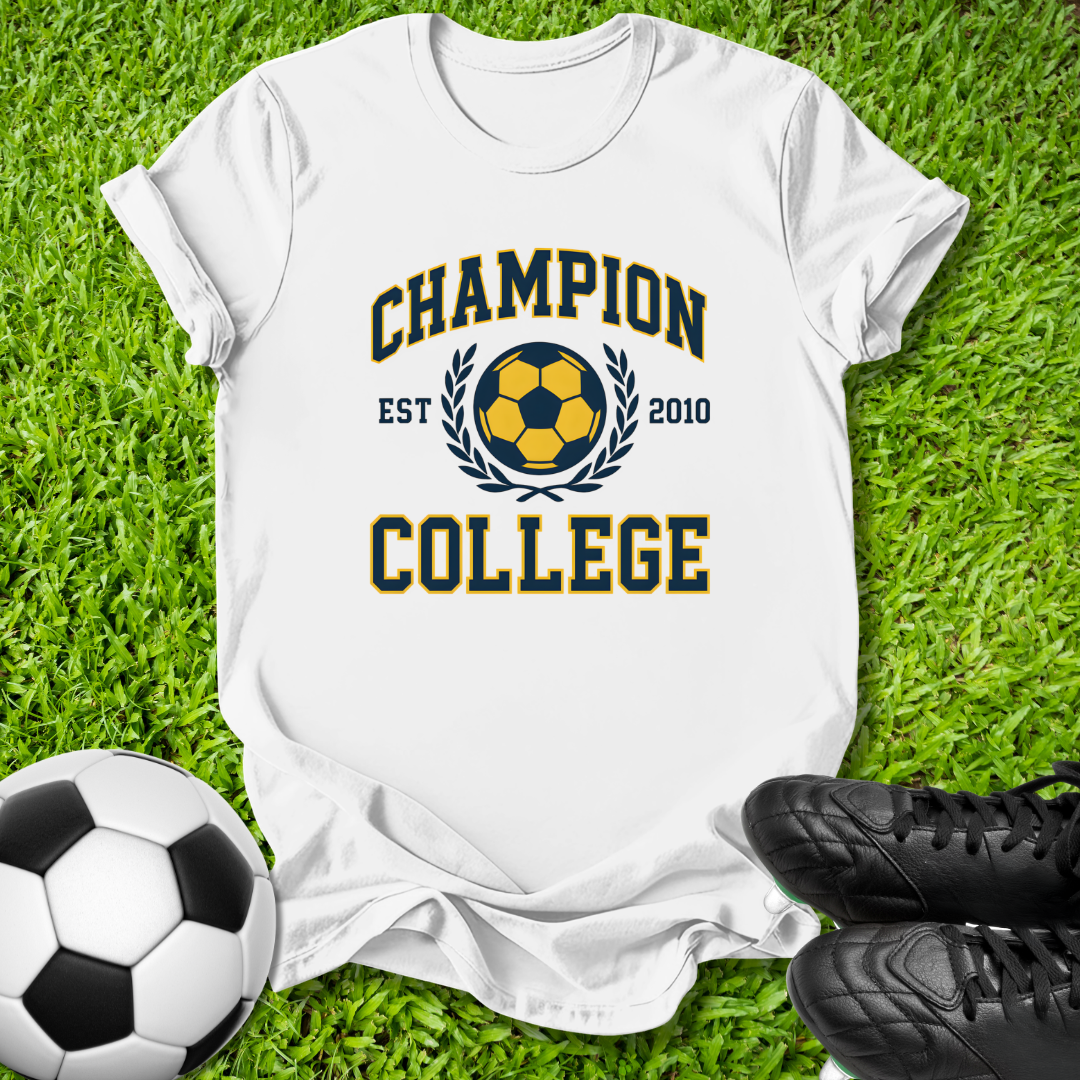 A white short-sleeve T-shirt laid flat on green grass, featuring a navy and yellow graphic that reads 'CHAMPION COLLEGE EST 2010' around a soccer ball motif. A soccer ball and black cleats are placed near the shirt as styling props.