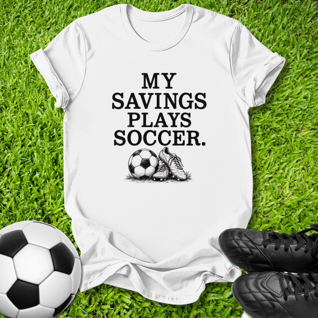 A white short-sleeve graphic T-shirt laid flat on green grass, with black text reading 'MY SAVINGS PLAYS SOCCER.' and a small soccer ball and cleat illustration.