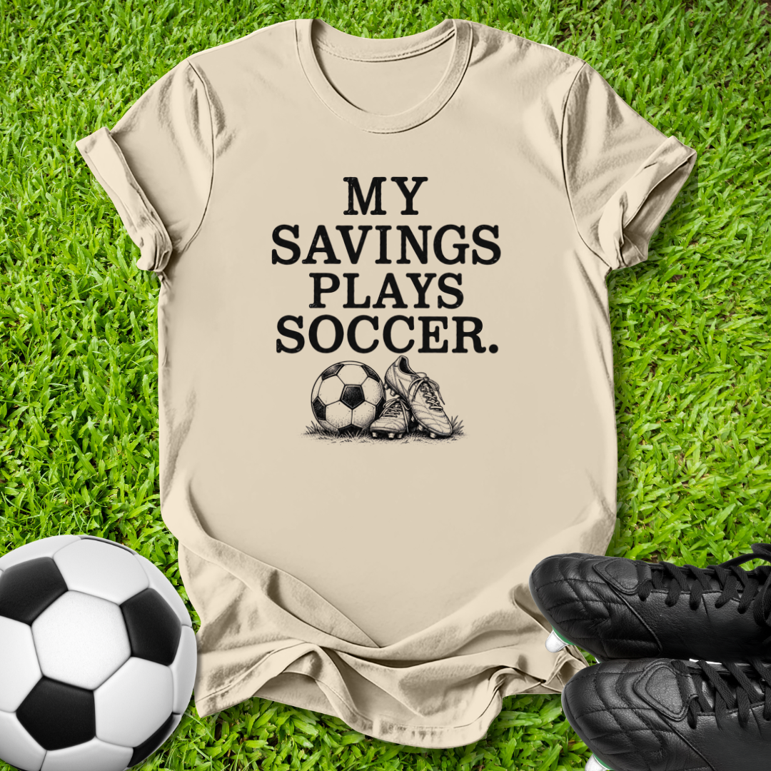 A beige graphic t-shirt laid flat on green grass, featuring the black printed text 'MY SAVINGS PLAYS SOCCER.' with a soccer ball and cleat illustration.