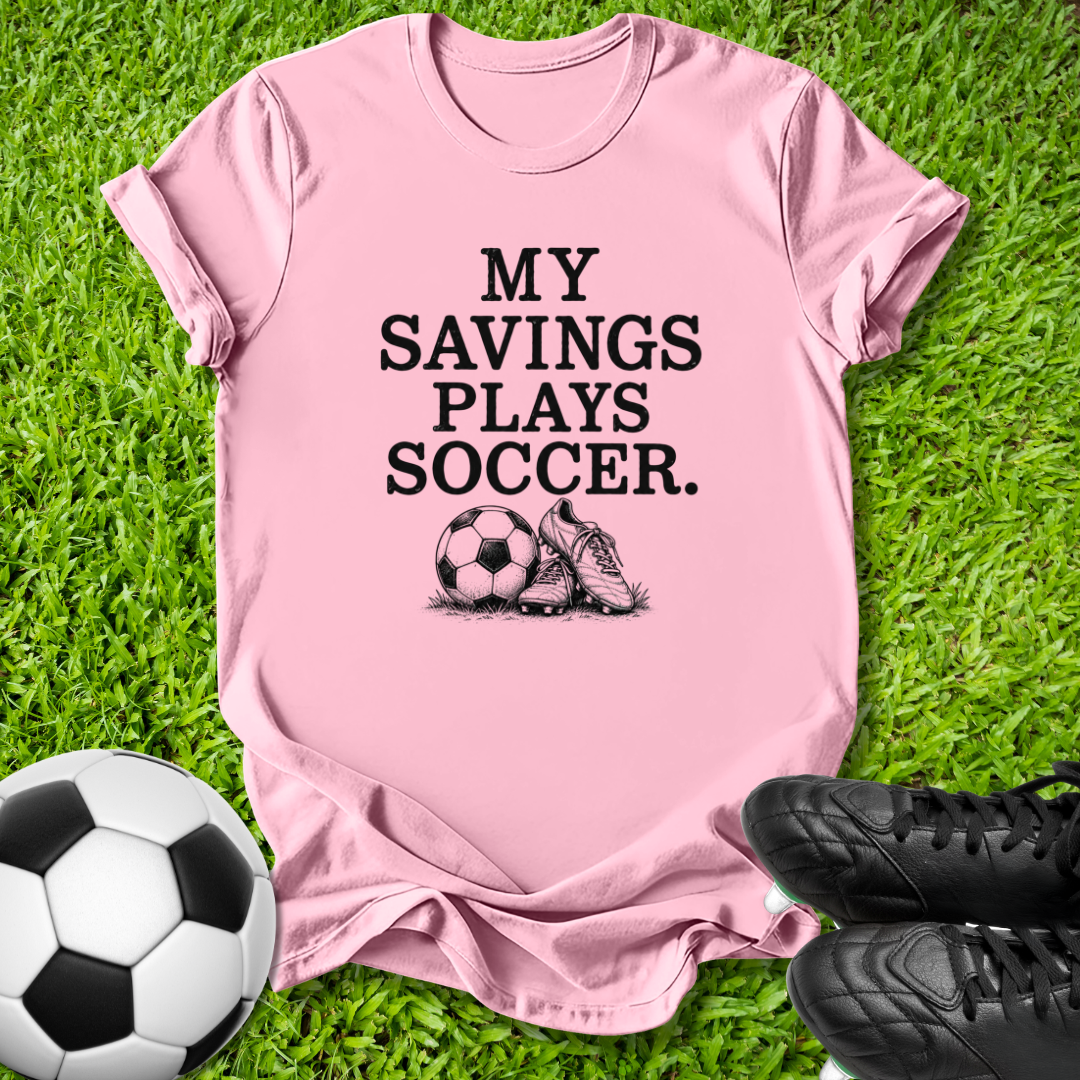 A pink short-sleeve t-shirt with the black text 'MY SAVINGS PLAYS SOCCER.' and a soccer ball and cleat graphic, laid flat on green grass with a soccer ball and black cleats as props.