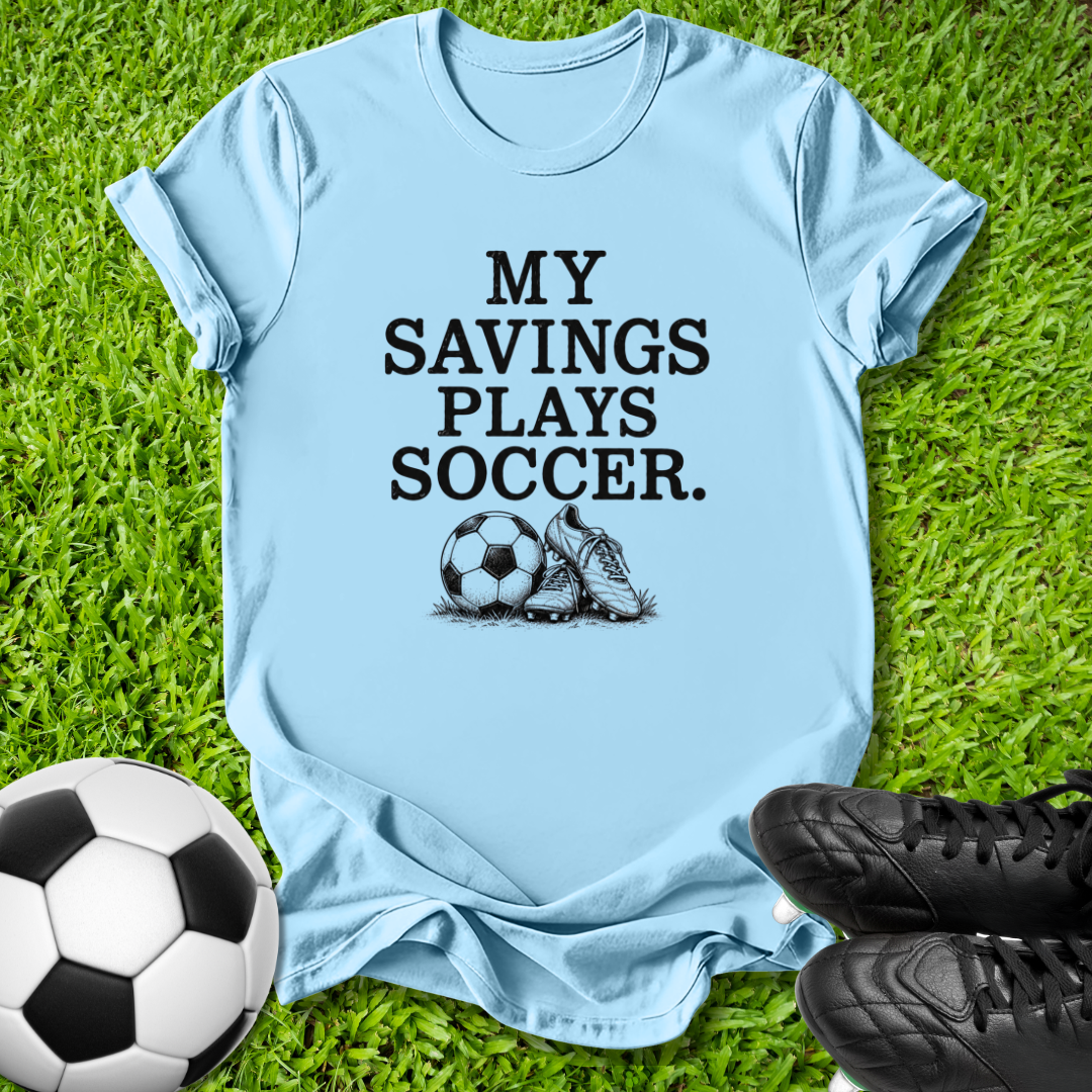 A light blue graphic t-shirt laid flat on green grass, featuring the black text 'MY SAVINGS PLAYS SOCCER.' above a soccer ball and cleat graphic.