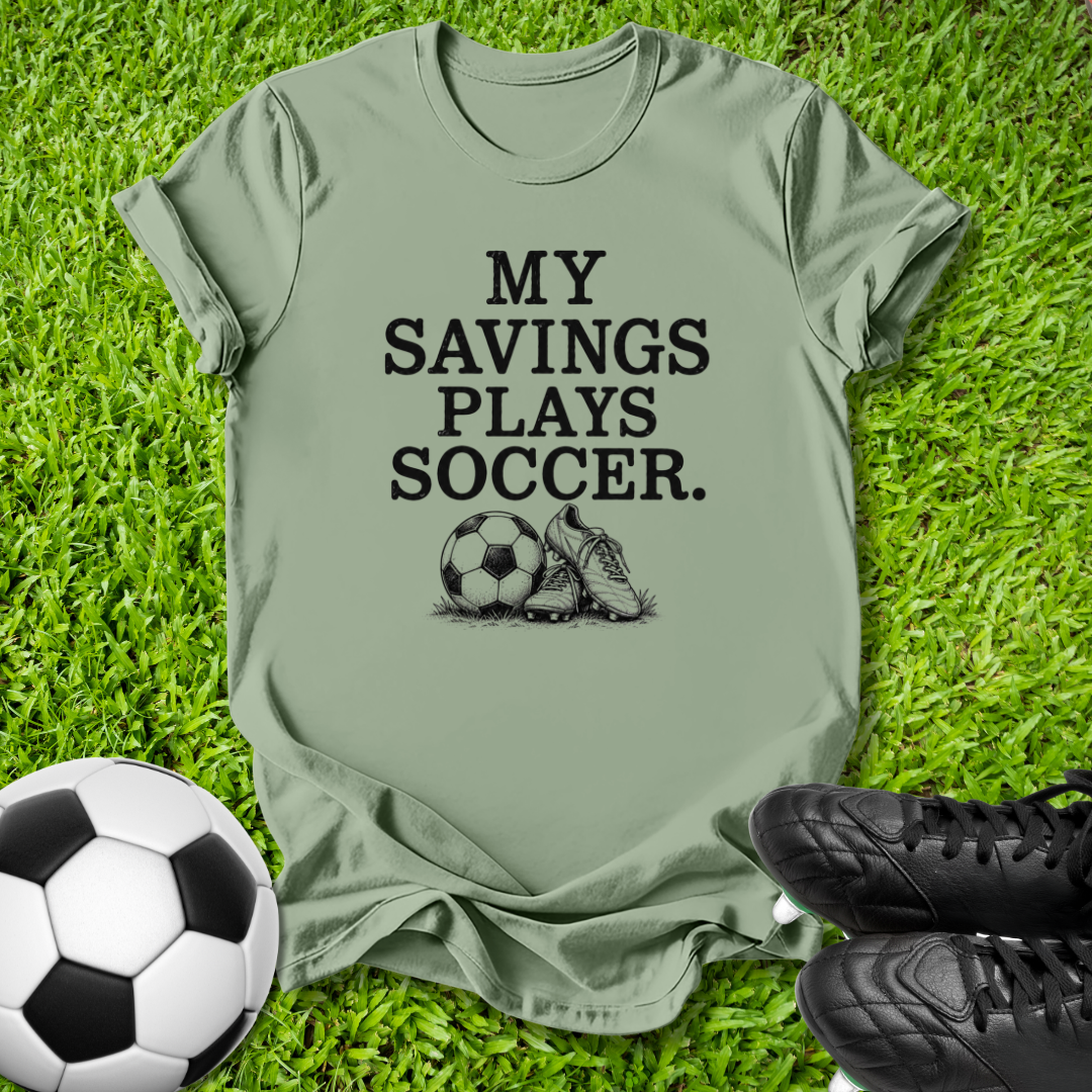 An olive green t-shirt with the black printed text 'MY SAVINGS PLAYS SOCCER.' and a soccer ball and cleat graphic, laid flat on green grass with a soccer ball and black cleats as props.