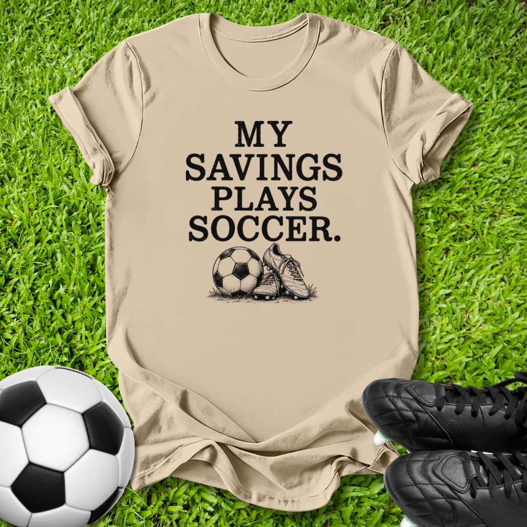 A beige graphic t-shirt laid flat on green grass, featuring the black text 'MY SAVINGS PLAYS SOCCER.' above a soccer ball and cleat graphic.