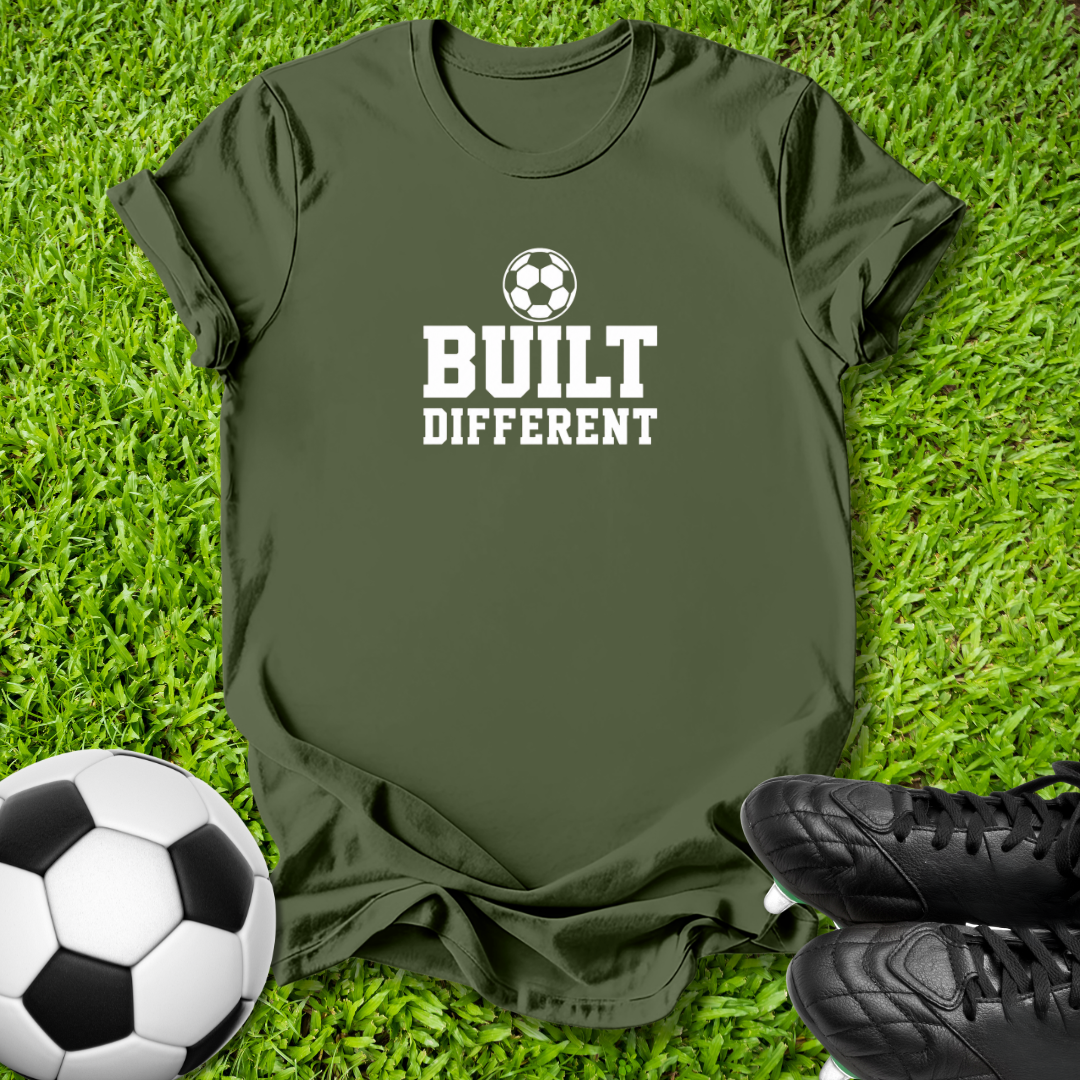 An olive green t-shirt with a white graphic reading 'BUILT DIFFERENT' and a soccer ball icon, laid flat on green grass with a soccer ball and black cleats nearby.