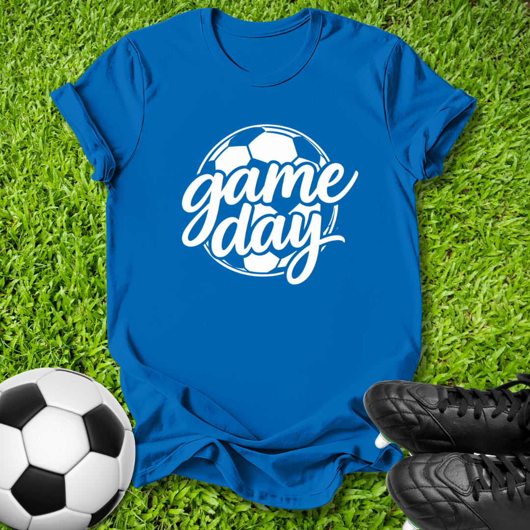 A royal blue short-sleeve t-shirt laid flat on green grass, featuring a white graphic that reads 'game day' over a soccer ball design. A soccer ball and black cleats are placed around it as props.