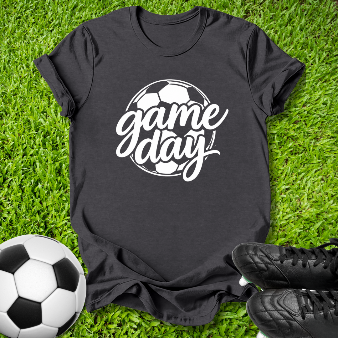 A black short-sleeve t-shirt with a white 'game day' soccer graphic, laid flat on green grass with a soccer ball and black cleats as props.