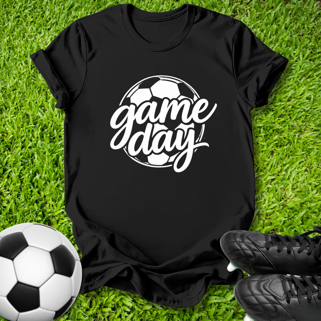 A black short-sleeve t-shirt with a white 'game day' soccer graphic, laid flat on green grass with a soccer ball and black cleats as props.