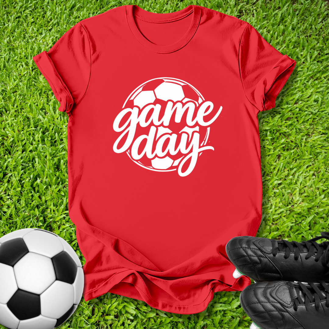 A red short-sleeve t-shirt with a white 'game day' soccer graphic, laid flat on green grass with a soccer ball and black cleats as props.