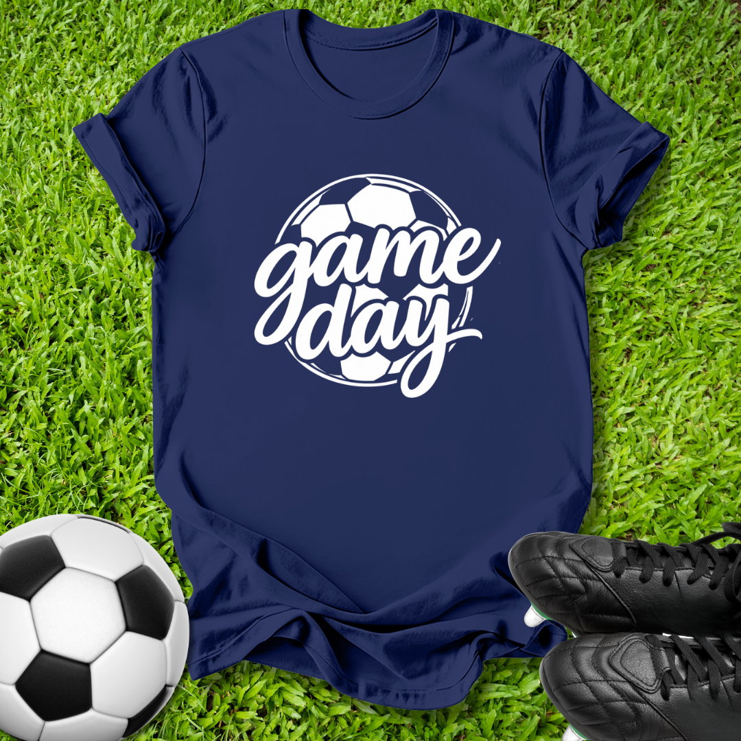 A navy blue short-sleeve t-shirt with a white 'game day' soccer graphic, laid flat on green grass with a soccer ball and black cleats as props.