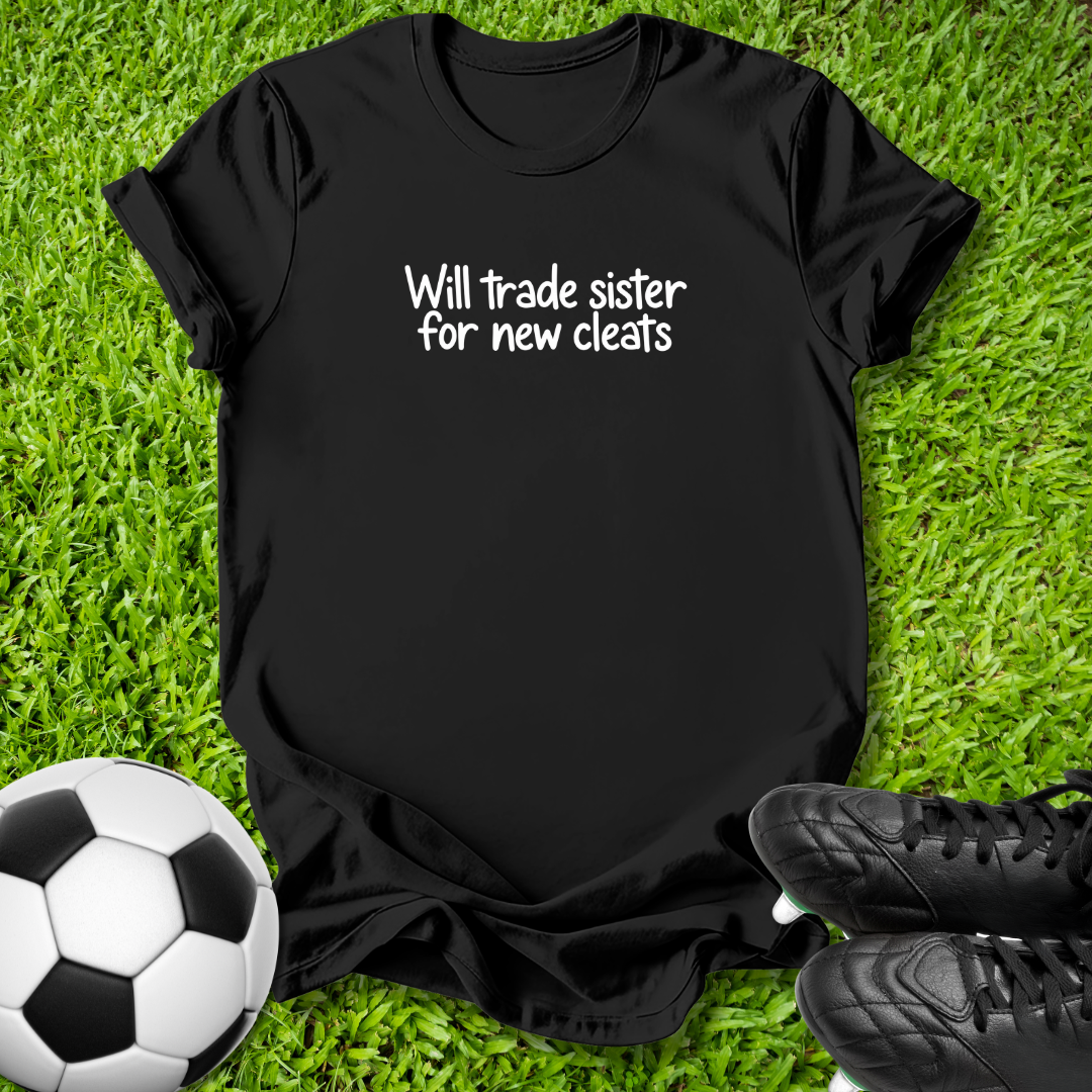 A black short-sleeve t-shirt with white text reading 'Will trade sister for new cleats' is laid flat on green grass, styled with a soccer ball on the left and black cleats on the right.