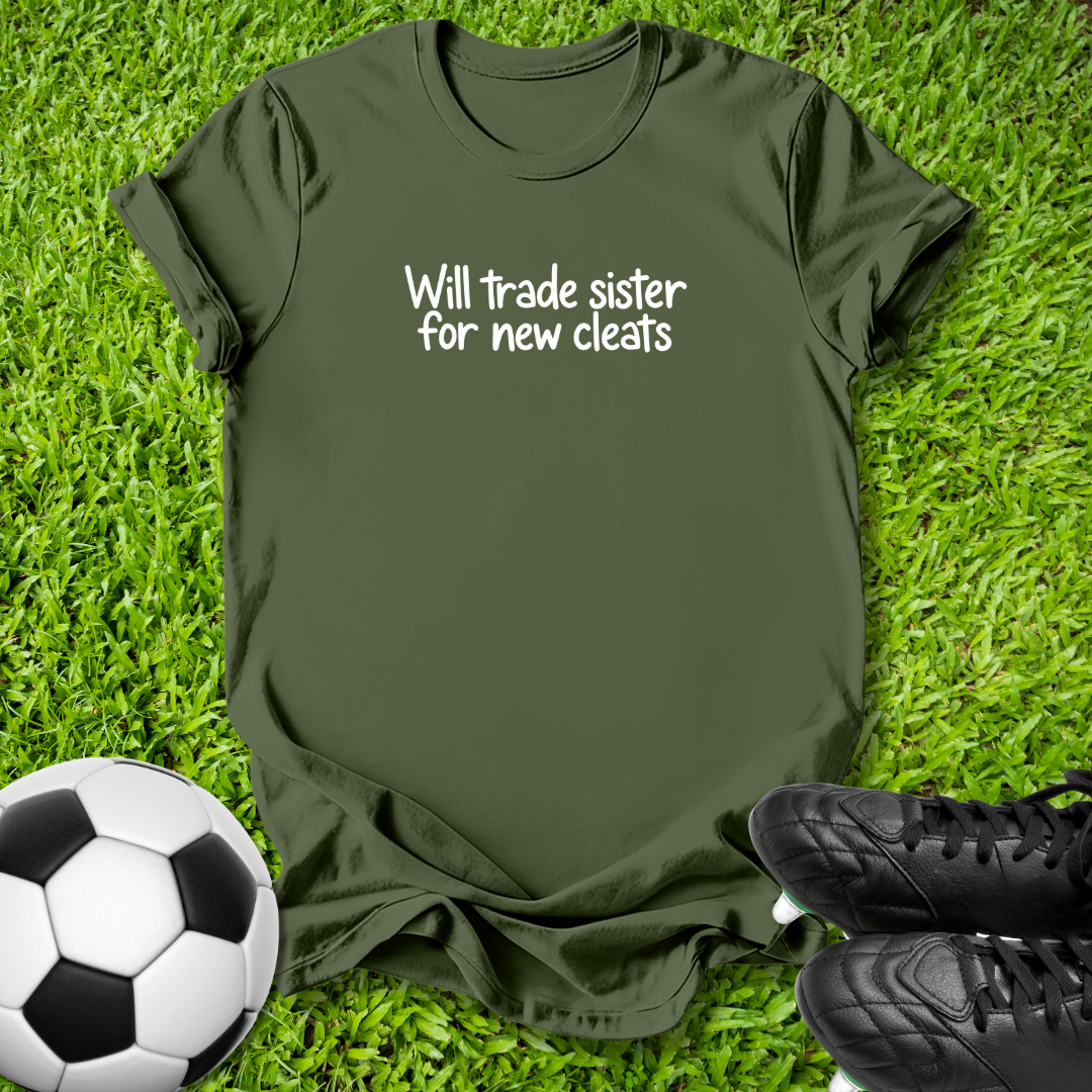 An olive green short-sleeve t-shirt with the white printed text 'Will trade sister for new cleats' laid flat on green grass, styled with a soccer ball on the left and black cleats on the right.