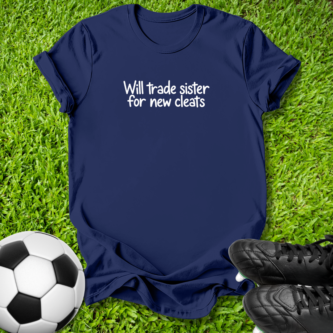 A navy blue short-sleeve t-shirt with the white text 'Will trade sister for new cleats' printed on the chest, laid flat on green grass with a soccer ball and black cleats as props.