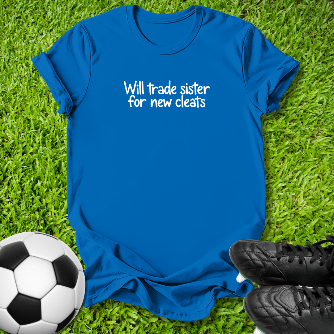 A royal blue short-sleeve t-shirt with white text reading 'Will trade sister for new cleats' laid flat on green grass, styled with a soccer ball on the left and black cleats on the right.