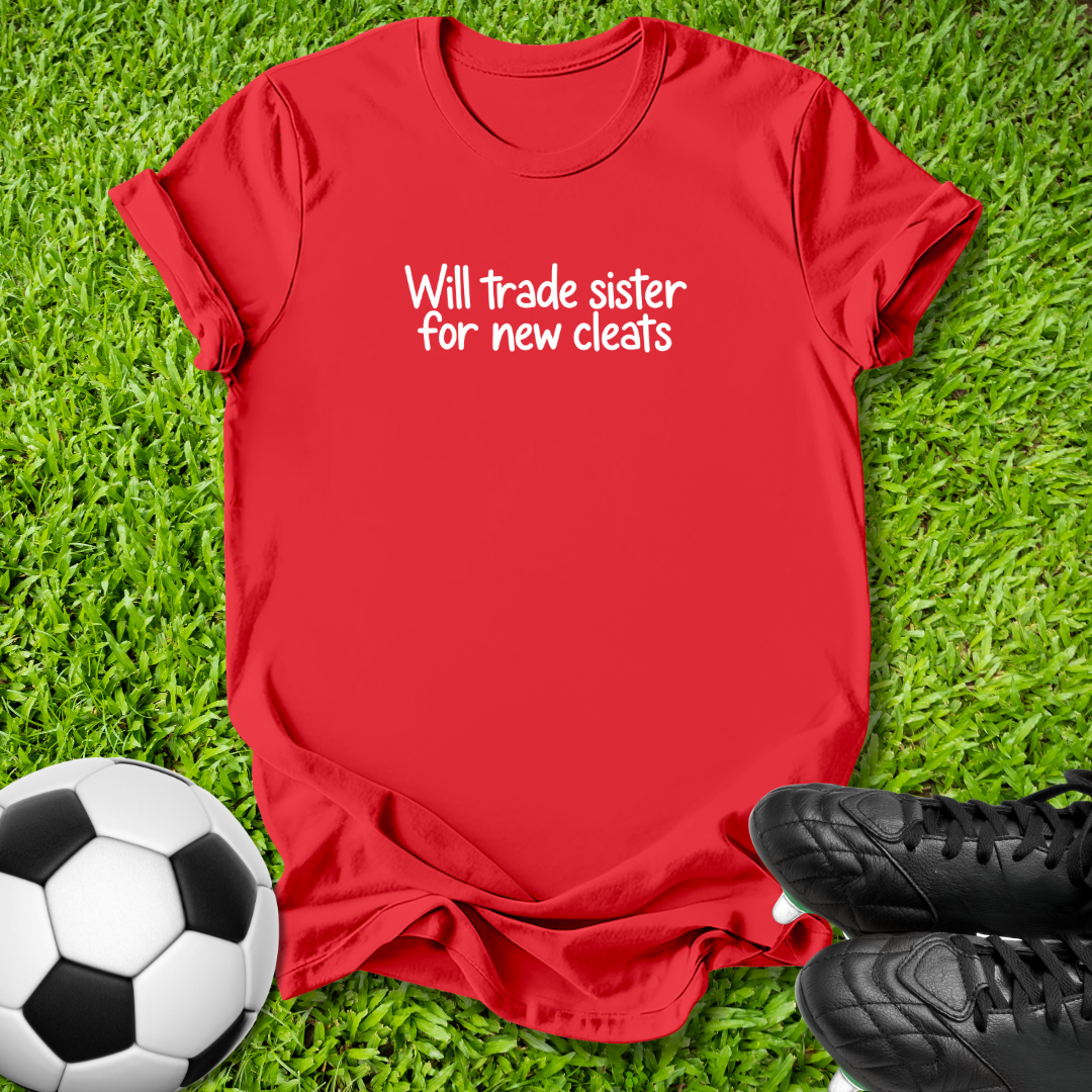 A red short-sleeve t-shirt with white text reading 'Will trade sister for new cleats' is laid flat on green grass, styled with a soccer ball on the left and black cleats on the right.