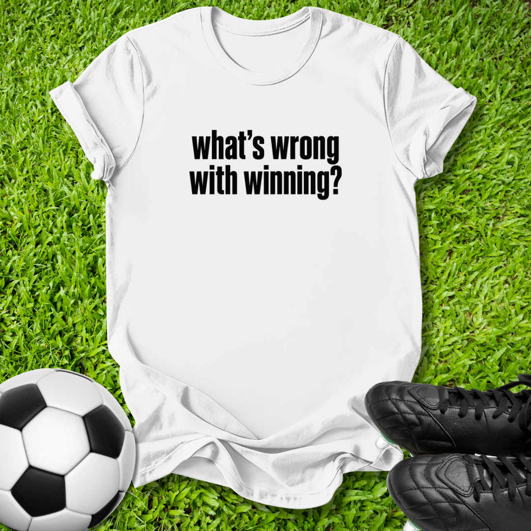 white  crewneck t-shirt with the text 'what's wrong with winning?' printed in black across the chest, laid flat on green grass with a soccer ball and black cleats as props.