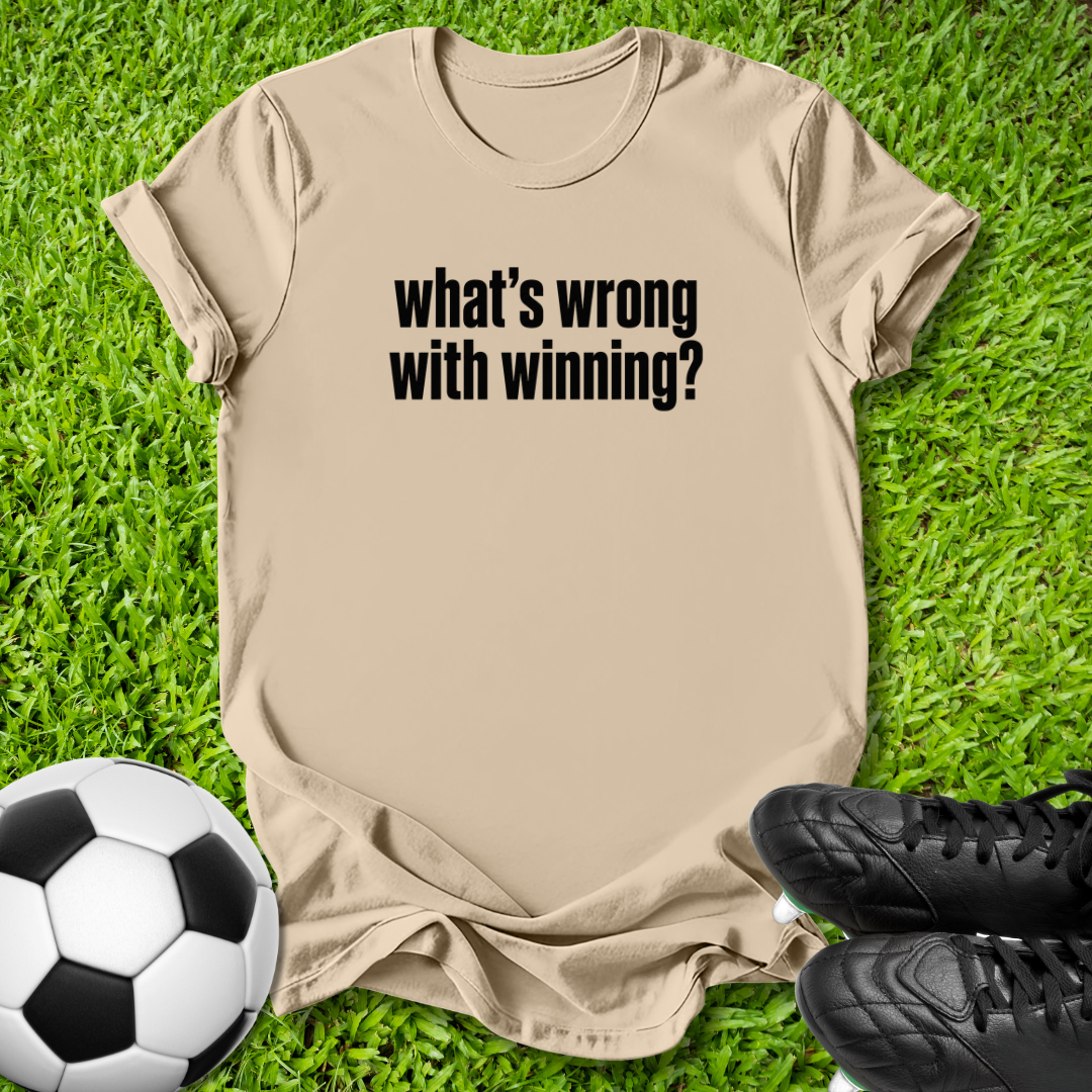 A beige graphic t-shirt with the text 'what's wrong with winning?' printed in black, laid flat on green grass with a soccer ball and black cleats as props.