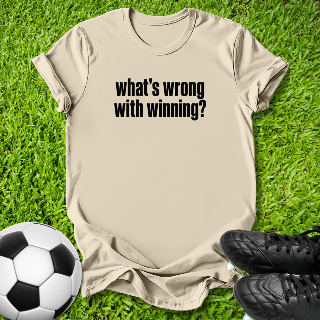 A beige graphic t-shirt with the text 'what's wrong with winning?' printed in black, laid flat on green grass with a soccer ball and black cleats as props.