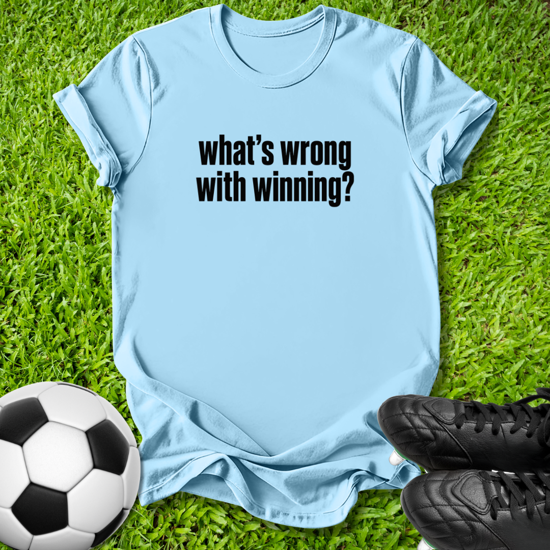 A light blue crewneck t-shirt with the text 'what's wrong with winning?' printed in black across the chest, laid flat on green grass with a soccer ball and black cleats as props.