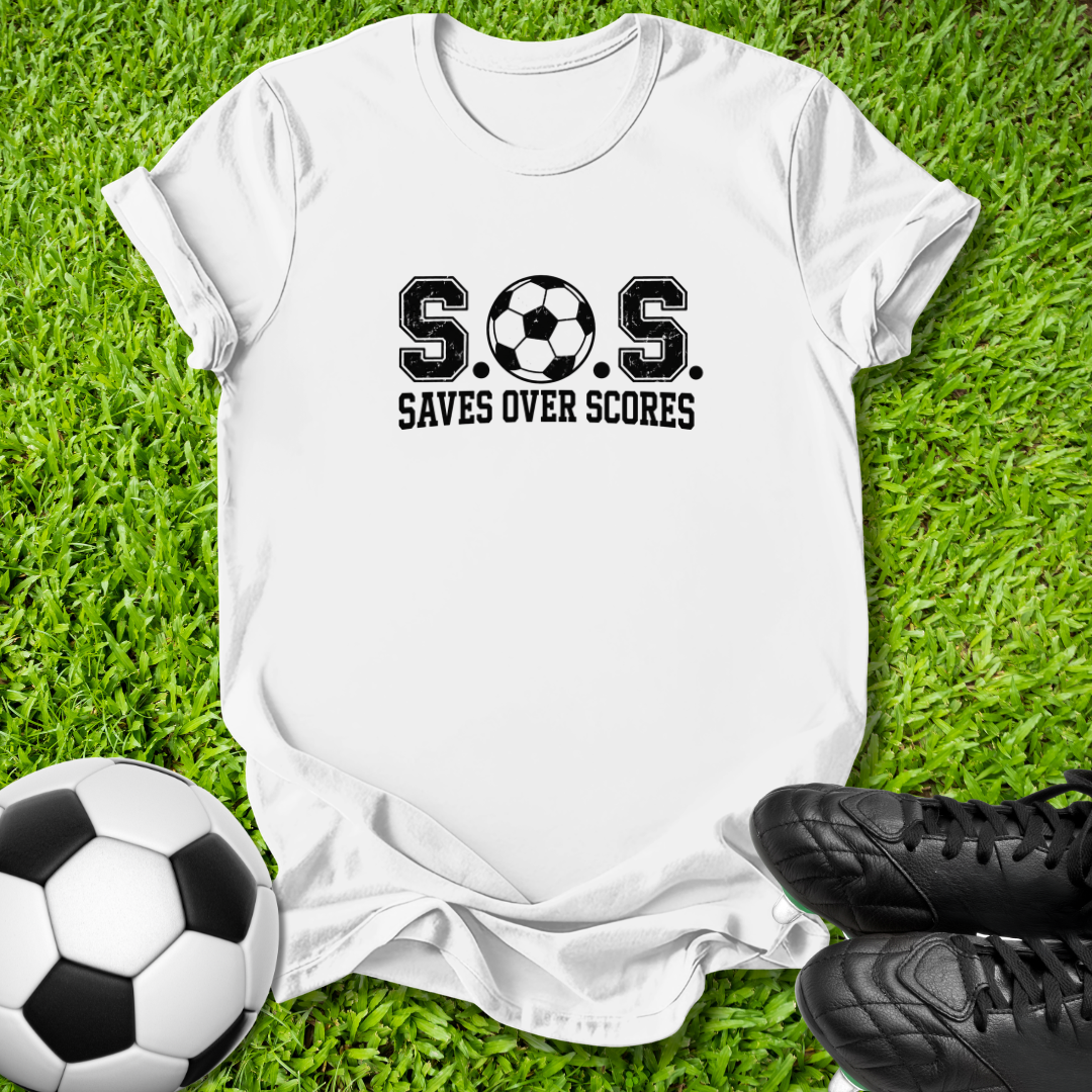 A white short-sleeve graphic T-shirt laid flat on green grass, with a soccer ball at the lower left and black cleats at the lower right. The shirt has a black printed slogan reading 'S.