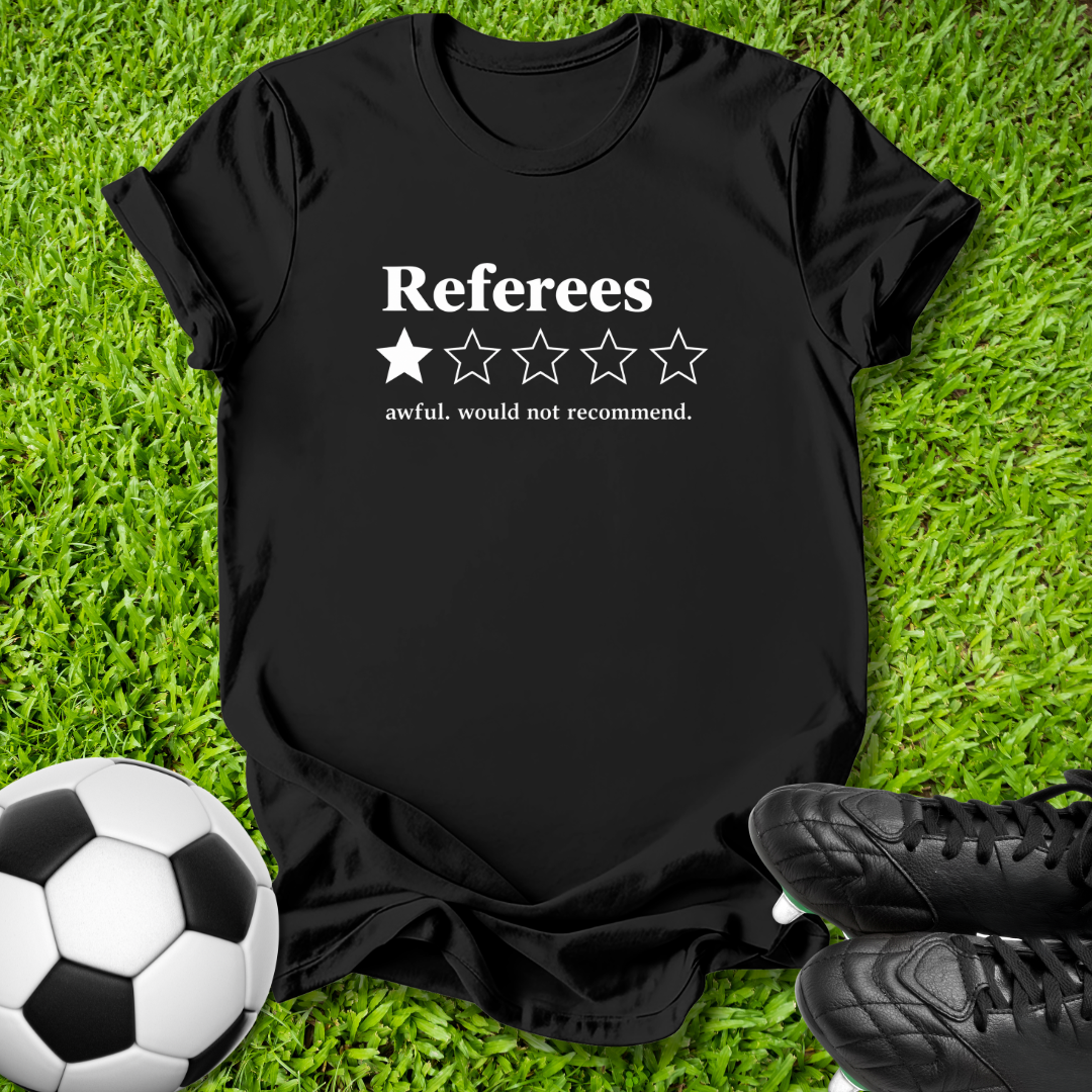 A black short-sleeve graphic T-shirt laid flat on green grass, with a soccer ball at the lower left and black cleats at the lower right. The shirt has white text reading "Referees" above a one-out-of-five star graphic and the phrase "awful, would not recommend.