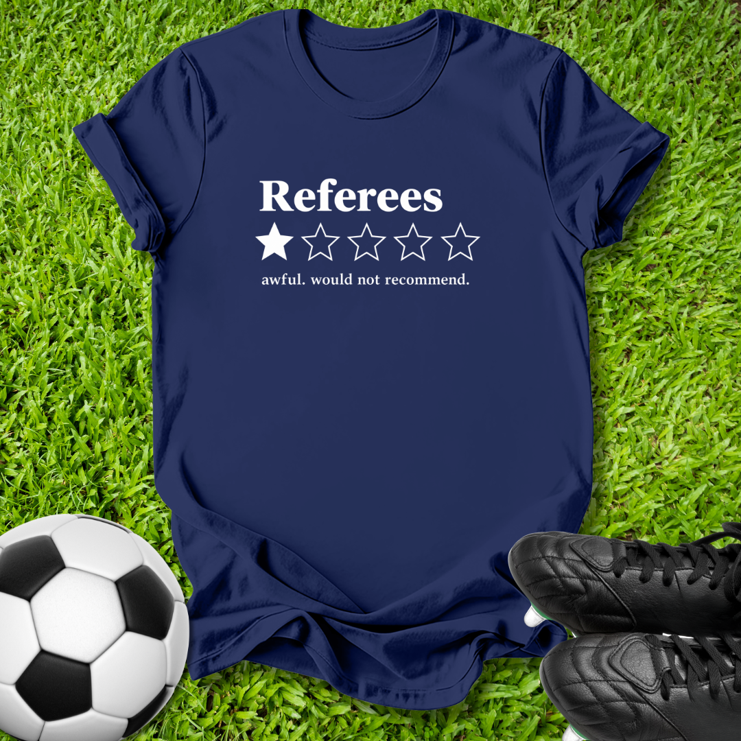 A navy blue graphic t-shirt laid flat on green grass, featuring a white printed design that reads 'Referees' above a one-star rating graphic and the phrase 'awful, would not recommend.' A soccer ball is visible in the lower left and black cleats in the lower right.