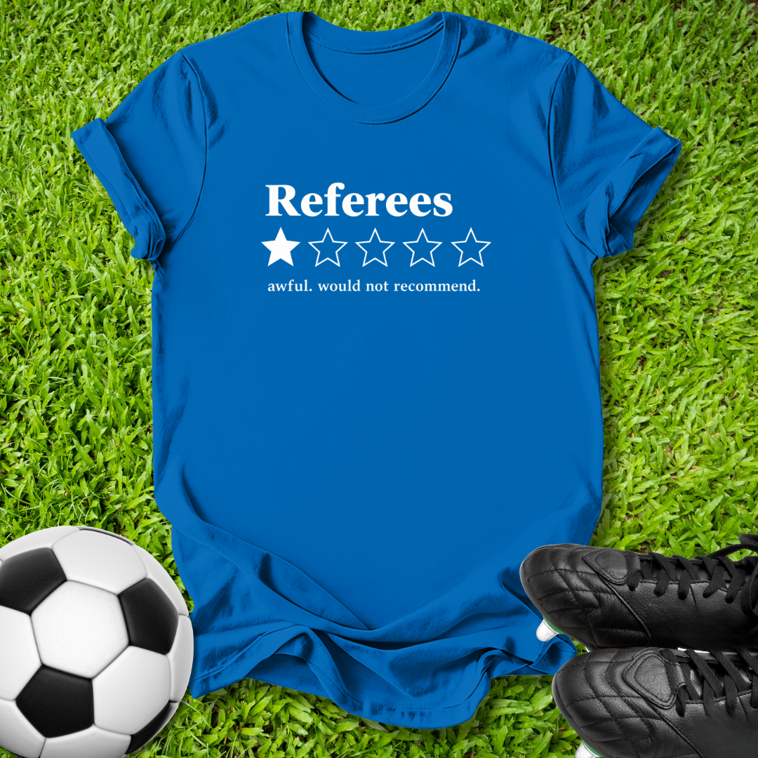 A royal blue short-sleeve t-shirt with a white graphic reading 'Referees' above a one-star rating, placed on green grass with a soccer ball and black cleats nearby.