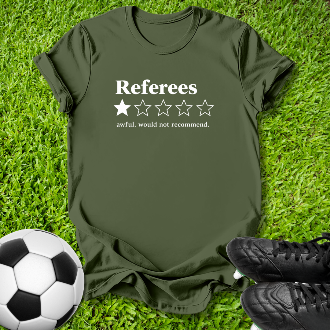 An olive green t-shirt with a white graphic reading 'Referees' and a one-star review design, laid flat on green grass with a soccer ball and black cleats as props.