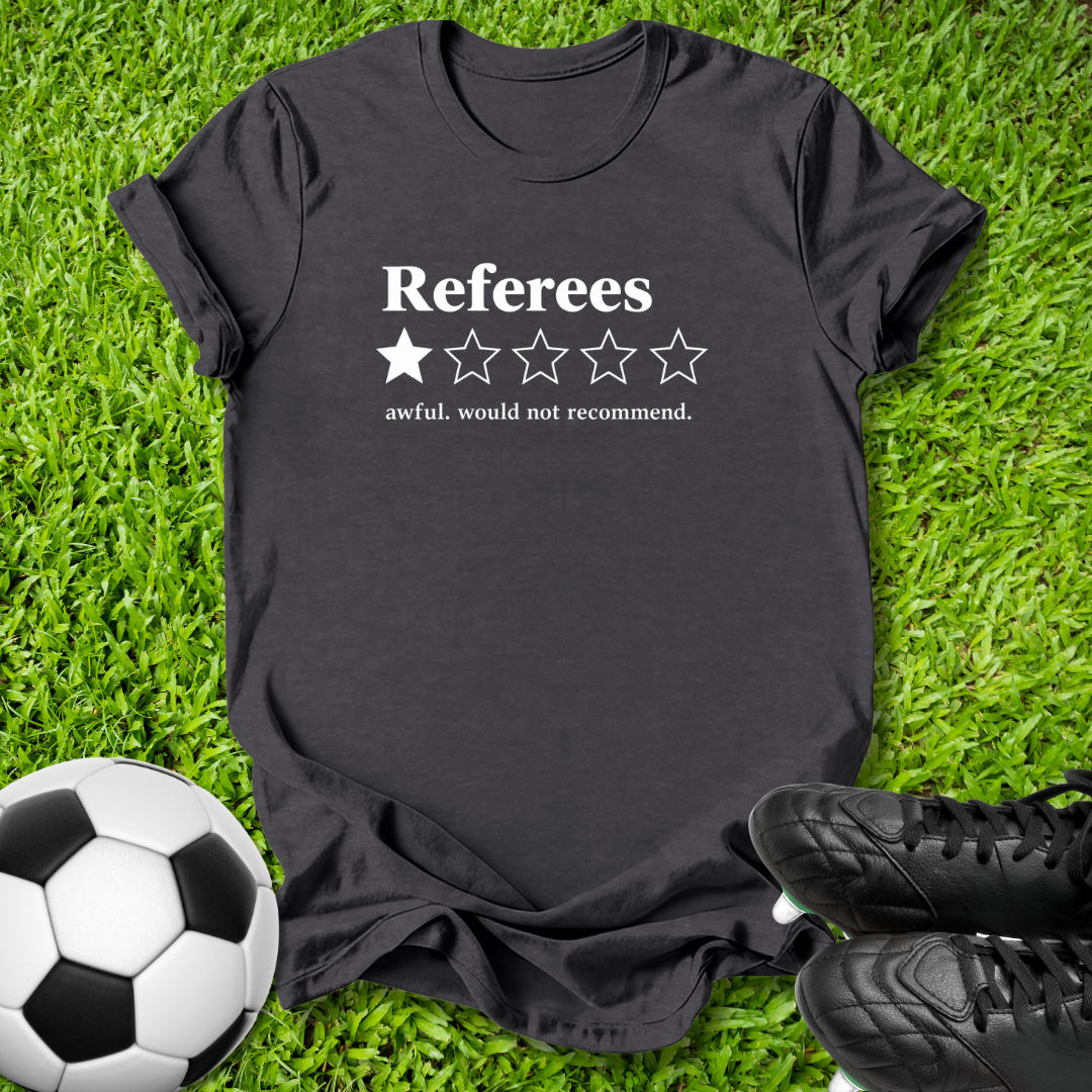 A black graphic t-shirt laid flat on green grass, featuring a white printed design that reads 'Referees' with one filled star and four empty stars, plus the phrase 'awful, would not recommend.' A soccer ball is visible at the bottom left and black cleats at the bottom right.