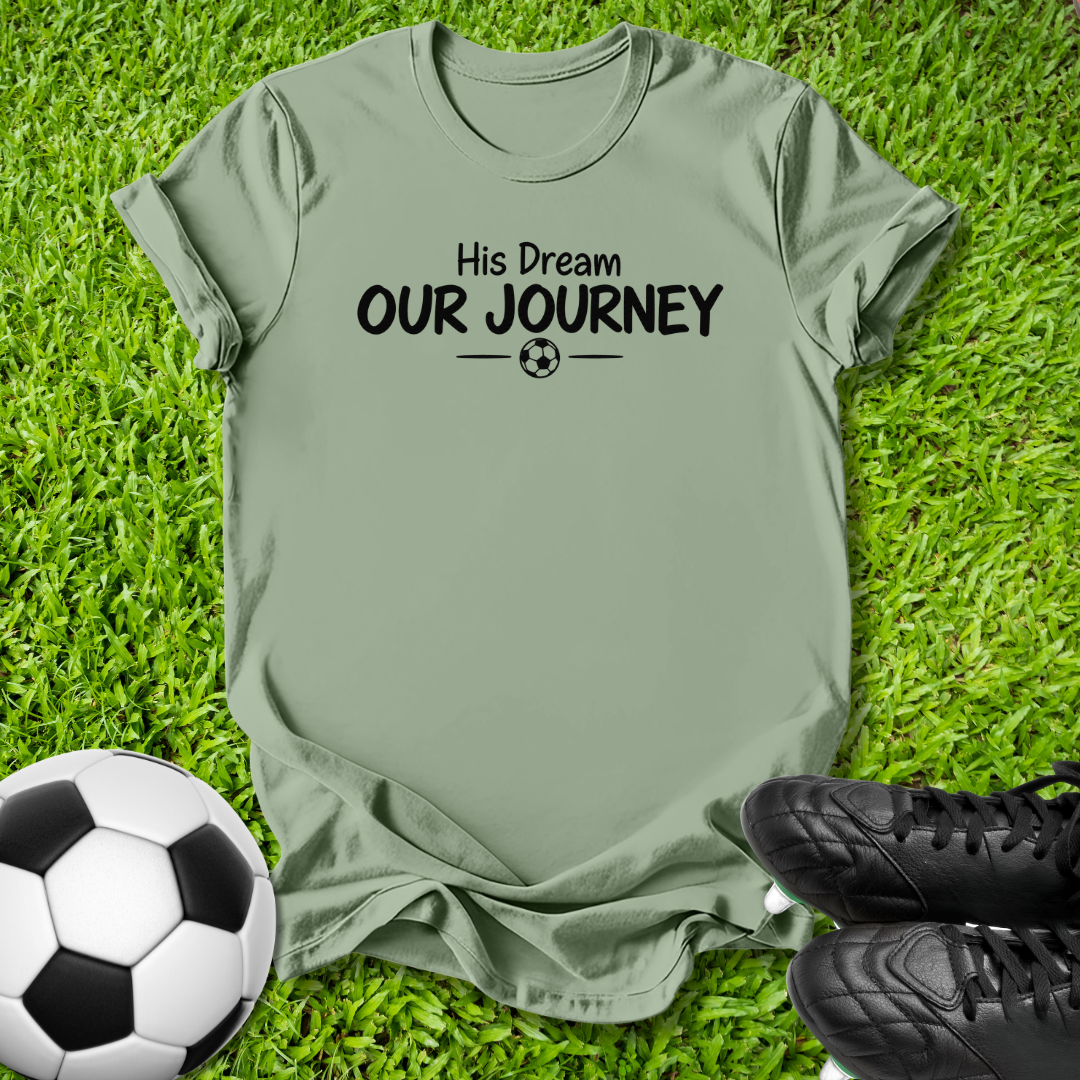 Green short-sleeve t-shirt with the black printed text 'His Dream OUR JOURNEY' and a small soccer ball graphic, laid flat on green grass with a soccer ball and black cleats nearby.