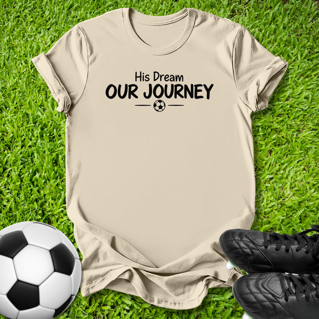 Natural short-sleeve t-shirt with the black printed text 'His Dream OUR JOURNEY' and a small soccer ball graphic, laid flat on green grass with a soccer ball and black cleats nearby.